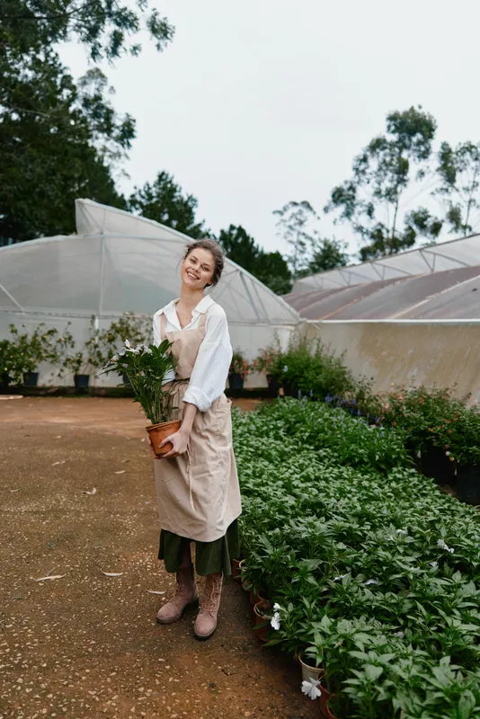 Young woman in an apron smiling while holding a potted flower in a greenhouse, surrounded by lush greenery and modern plant growing structures