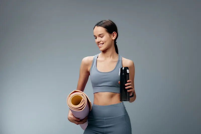 Fitness-focused woman in athletic wear stands with a rolled mat and a water bottle, ready for a workout. Grey studio backdrop highlights her athletic figure and health vibe.