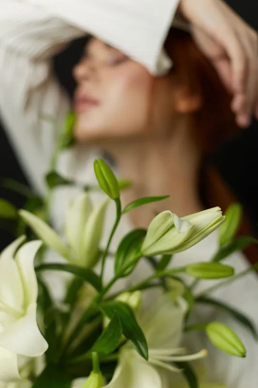 Elegant young woman with long hair holding a bouquet of white lilies, expressing a serene emotion against a dark background