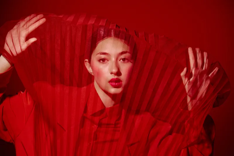 Portrait of a woman in vibrant red clothing and lips, holding a sheer fabric with hands visible behind it. Studio lighting highlights bold fashion and beauty concept.