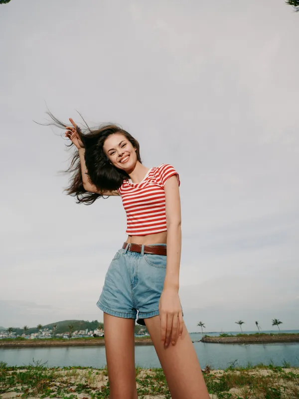 A carefree young woman stands on a sandy shoreline wearing a red striped crop top and denim shorts, wind lifting her hair, bright smile, relaxed pose against a clear sky.