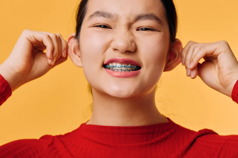 Teenage girl with braces smiling and pulling ears on orange background. Young female in red sweater with orthodontic treatment showing happiness and playful expression in studio.