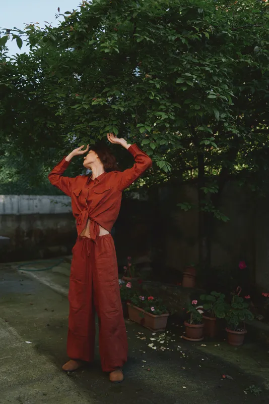 Cheerful woman outdoors wearing a red outfit, posing with arms raised, enjoying a sunlit garden scene, lush greenery and potted plants creating a relaxed lifestyle moment