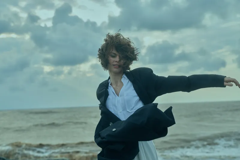 Windy beach motion with a blazer and dress, portraying confidence and professional style against the vast ocean, dramatic sky, and sweeping horizon during a windy day