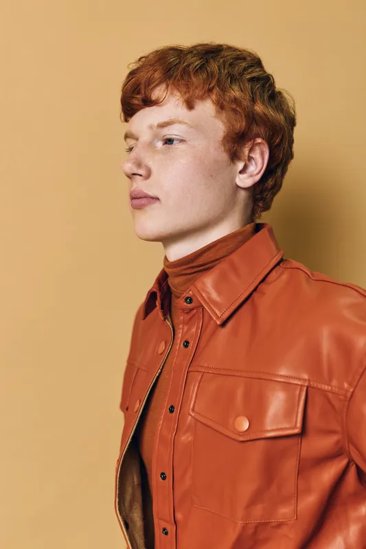 Young man with red hair wearing brown leather jacket and shirt posing against beige background. Profile portrait with serious expression and styled hair in studio lighting.