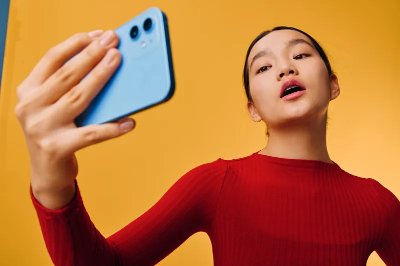 Woman with smartphone taking selfie indoors on yellow background wearing red sweater. Young adult female capturing self portrait with mobile phone in casual clothing for social media or communication.