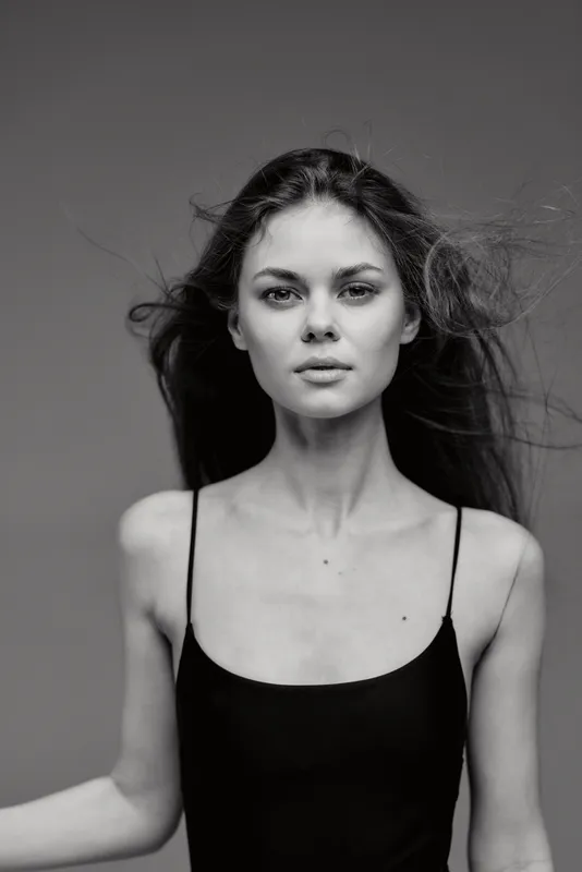 Black and white portrait of a young woman with flowing hair wearing a black sleeveless top, looking confidently at the camera.