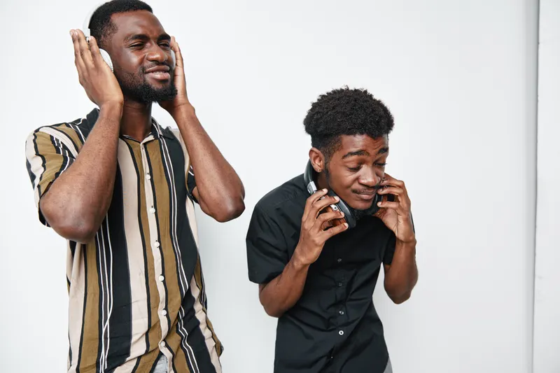 Two young black men with dark skin enjoying music with headphones, smiling and having fun together in casual clothes on white background.