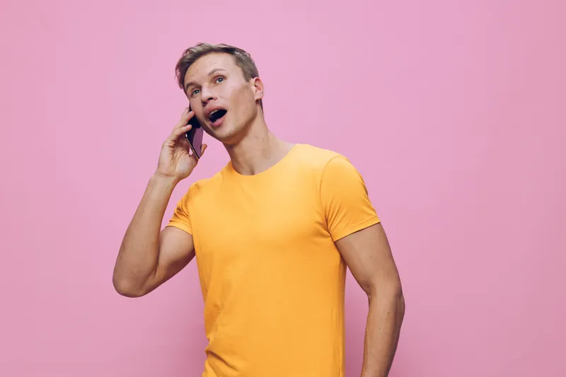 Smiling man talking on mobile phone wearing bright yellow t-shirt against pastel pink background, casual style, cheerful mood, indoor studio portrait, lifestyle concept, communication, modern fashion.