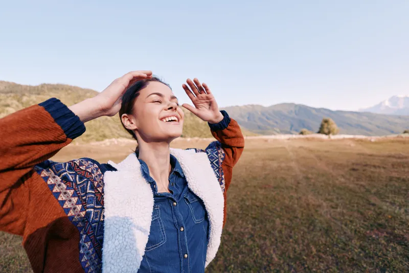 Woman smiling with eyes closed outdoors in countryside, hands raised enjoying nature and warm sunshine, happiness and freedom in casual jacket and denim, relaxed joyful portrait in meadow.