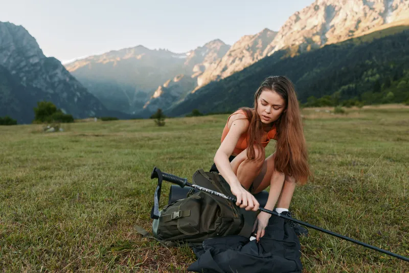 A young woman outdoors in alpine meadow, adjusting a trekking pole beside a backpack. The distant mountains glow under soft sunlight, creating a calm, adventurous outdoor atmosphere.