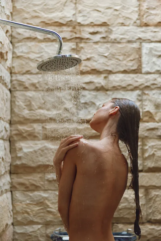 woman showering outdoors with long wet hair under rain showerhead, natural stone wall, serene and refreshing moment, morning routine relaxation
