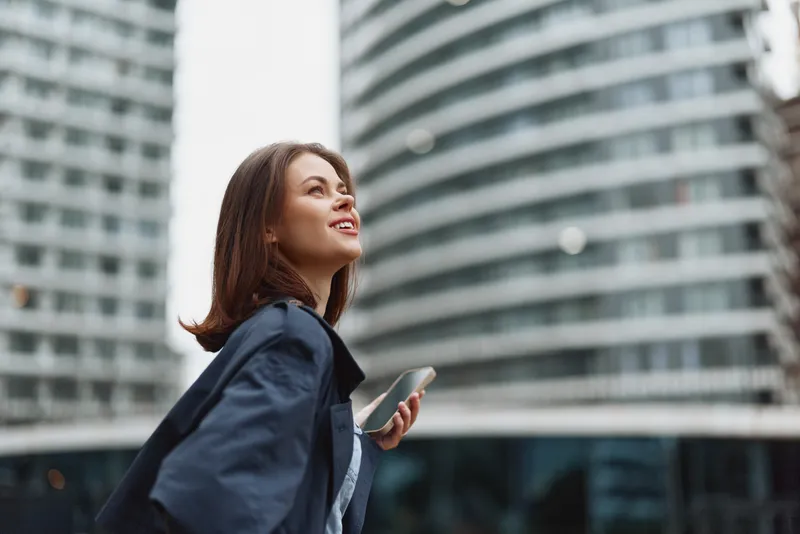 A confident young businesswoman in a dark blazer looks up while holding a smartphone in an urban environment with modern glass buildings.