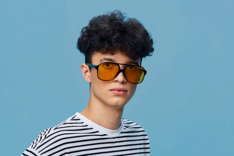 Young man with curly hair wearing yellow sunglasses and a striped shirt, posing against a smooth blue background with a casual serious expression.
