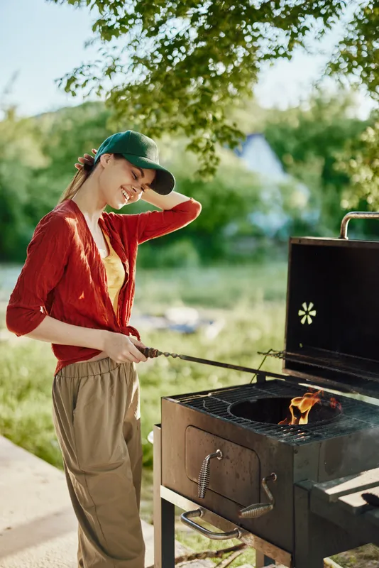 Young woman grilling outdoors in summer, wearing casual clothing and a cap, enjoying the moment with a smile, surrounded by greenery and a clear blue sky