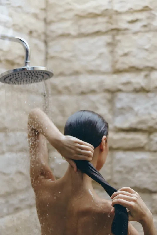 Woman washing hair under outdoor shower with flowing water against textured stone wall, natural light highlighting wet skin and dark hair strands.
