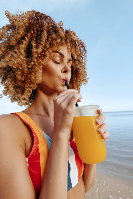 Woman with curly hair sips orange drink through straw, holding a mug near the beach, ocean and blue sky in background, summer relaxation, hydration, sunlight, seaside vacation mood
