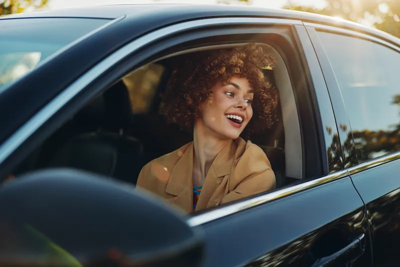 A smiling woman sits in a car window, enjoying a sunny drive with retro film color. Warm tones and soft light create a candid lifestyle moment of joy and everyday travel.