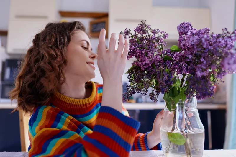 A woman in a colorful striped sweater sits at a table in a cozy home, admiring purple flowers in a glass vase. A bright, calm moment of everyday happiness and mindful living.
