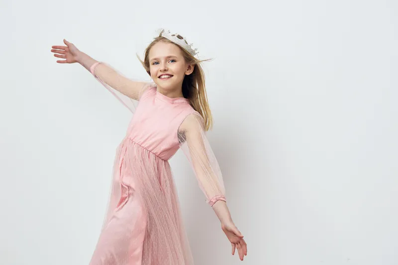 Joyful girl in a pink dress with a crown, expressing happiness against a light background, perfect for seasonal themes like spring or celebrations