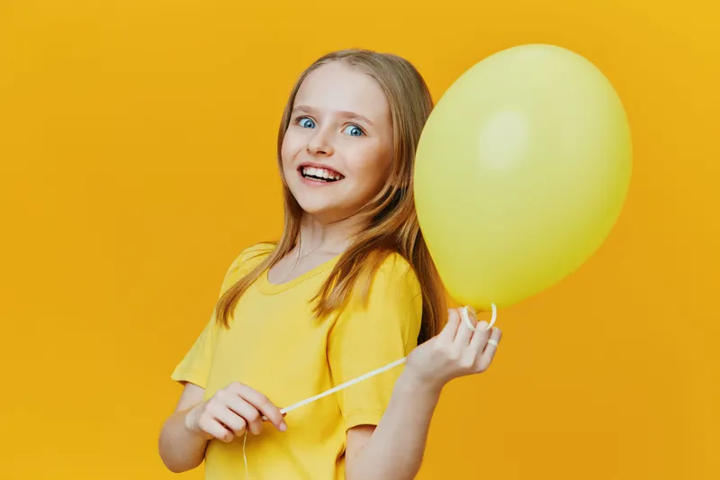 Happy girl in a yellow shirt holding a yellow balloon against a bright yellow background, expressing joy and fun, perfect for celebrations or children s events