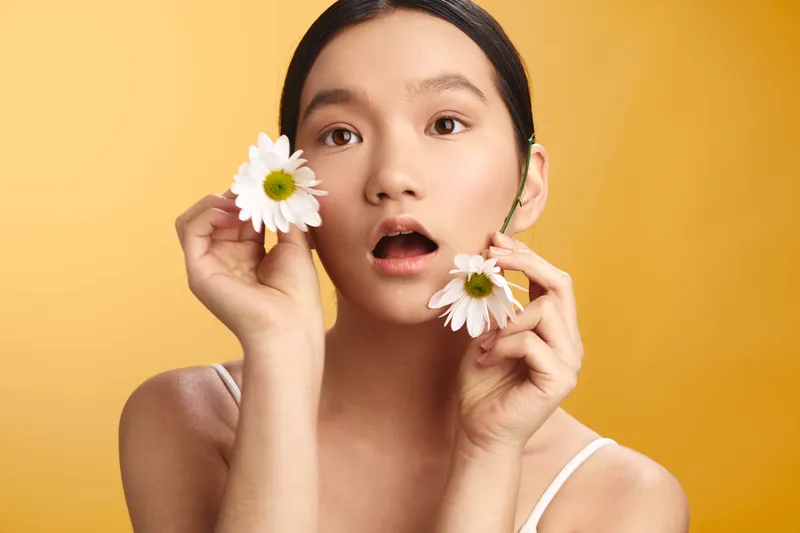 asian woman with a surprised expression holding white daisies near her face against a warm yellow studio backdrop, creating a fresh, playful mood and a striking, contemporary portrait.