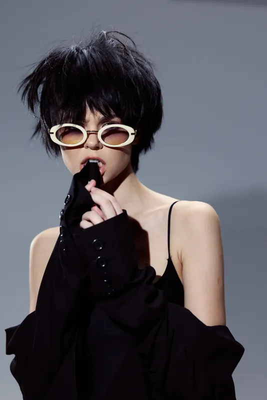 Stylish woman with short black hair wearing white sunglasses and black attire in a modern fashion portrait.