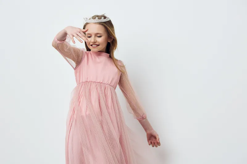 Charming young girl in a pink tulle dress, joyfully posing with a crown against a minimalist white background, capturing a playful and whimsical mood for children s fashion photography