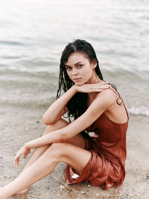 Young woman sitting on the beach with wet hair wearing a brown dress, looking thoughtfully by the sea shore in a serene summer setting.