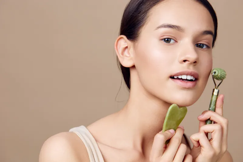 Young woman using jade roller and gua sha for skincare on neutral background, promoting healthy skin and beauty routines