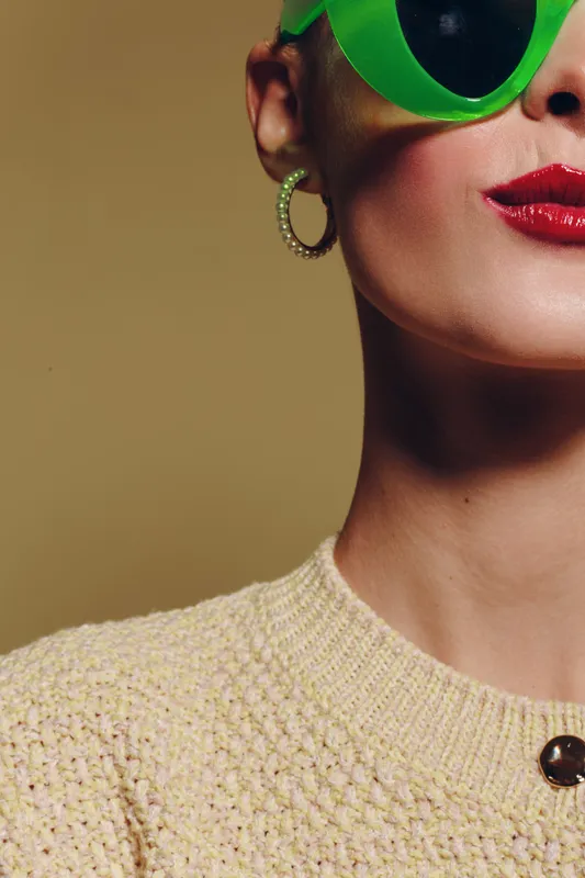 Close-up of a woman wearing green sunglasses and red lipstick, dressed in a beige knitted sweater with hoop earrings. Fashion concept with bold makeup and accessories.