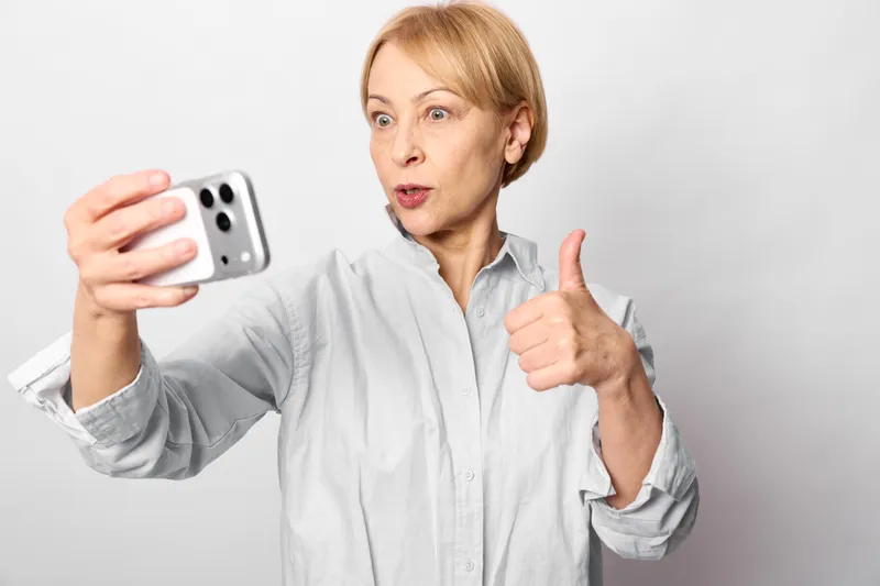 Woman holding smartphone, thumb gesture, selfie pose, surprised expression, technology concept, portrait on white background, studio lighting, neutral mood