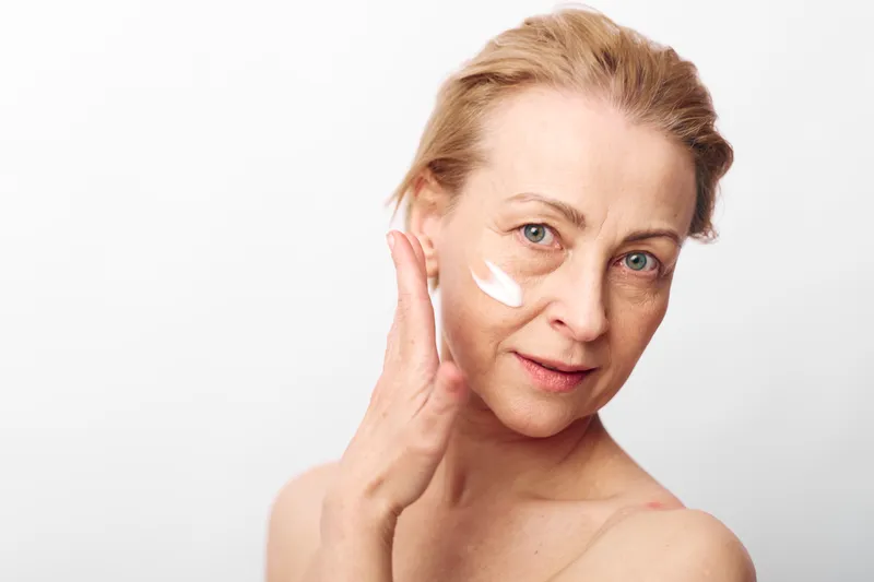 Mature woman with light skin applies moisturizing cream on face with hand, looking at camera on white background. Concept of skincare, aging beauty, self-care, and natural wellness.