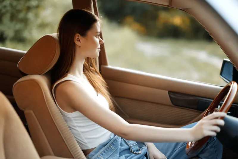 Young woman sitting in a vintage car, reflecting on life, wearing a casual white tank top and denim jeans, with a serene expression in a natural outdoor setting.