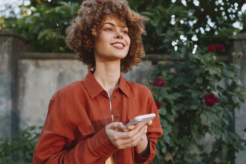 A smiling woman with curly hair outdoors holds a phone, wearing a casual red shirt. Garden and greenery in the background create a natural, warm mood focused on lifestyle and everyday moments