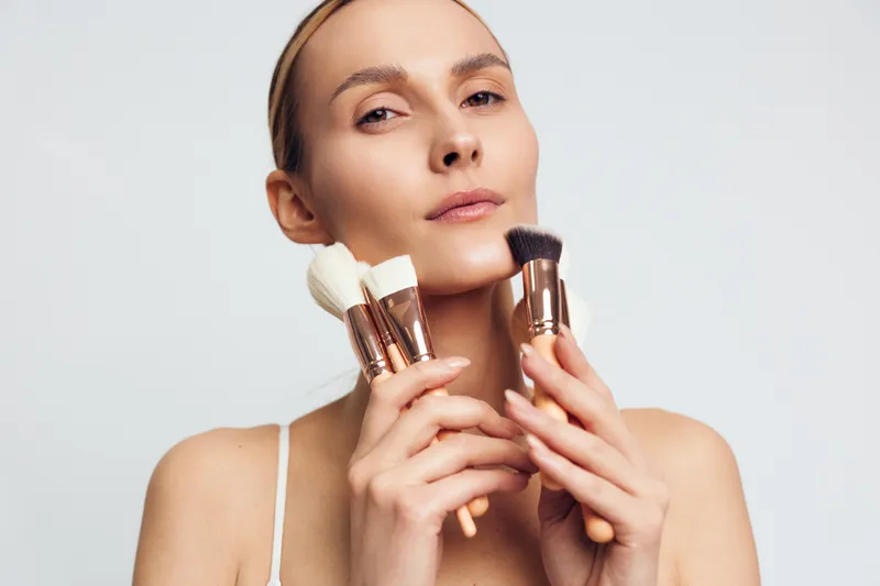 Young woman holding makeup brushes close to her face with neutral expression. Studio portrait on white background. Concept of beauty tools, cosmetics, and skincare routine.