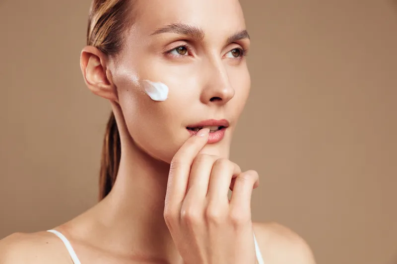 Young woman applies moisturizing cream on face looks thoughtful with natural skin care routine isolated on beige background closeup portrait with soft lighting and calm expression