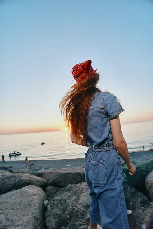 lifestyle woman in modern boho-western denim street style with red bandana and flowing hair at seaside during sunset, film color tones creating warm mood