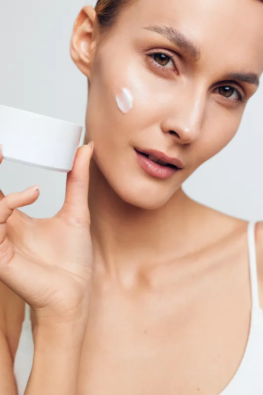 Young woman with clear skin applies moisturizing cream on her cheek holds white jar close to face has natural makeup and gentle expression isolated on light background