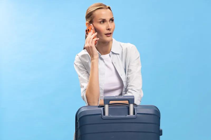 Young woman in white shirt holds phone to ear and leans on large blue suitcase against light blue background. Concept of travel, communication, waiting, and trip planning.
