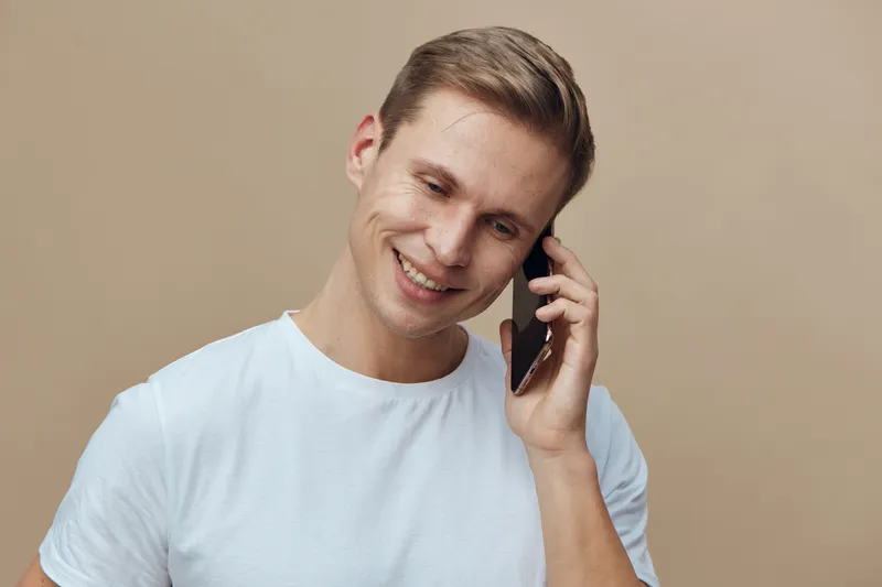 Smiling young man talking on mobile phone with friendly expression, casual white t-shirt, light beige background, studio portrait, people lifestyle concept, communication, happiness, leisure, technolo