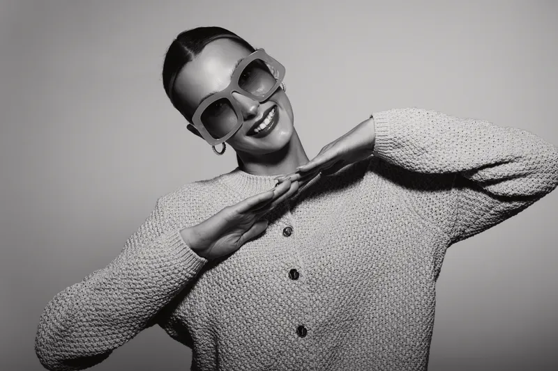 Stylish smiling young caucasian woman with fair skin wearing sunglasses and knitted sweater poses isolated on grey background.