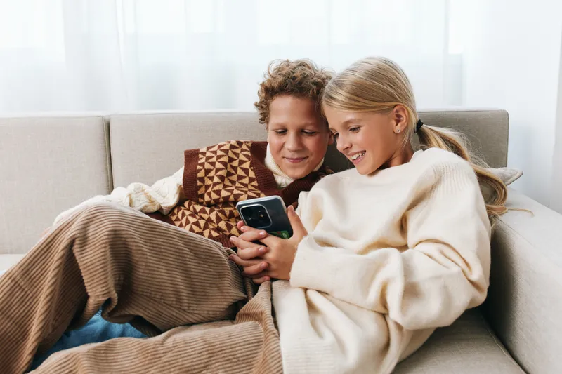 Two smiling children sit close on a soft sofa, sharing a mobile device. Cozy sweaters, warm light, and relaxed mood convey gentle family bonding and comfort.