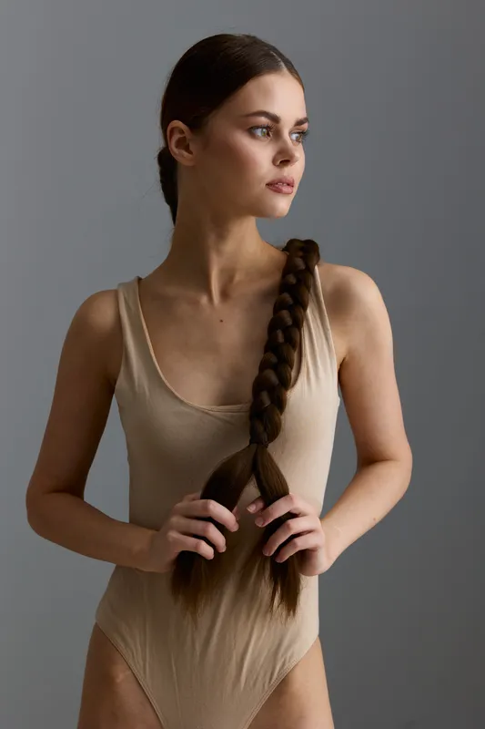 young woman with long braided hair wearing beige bodysuit standing against gray background, natural light, minimalistic style, thoughtful expression