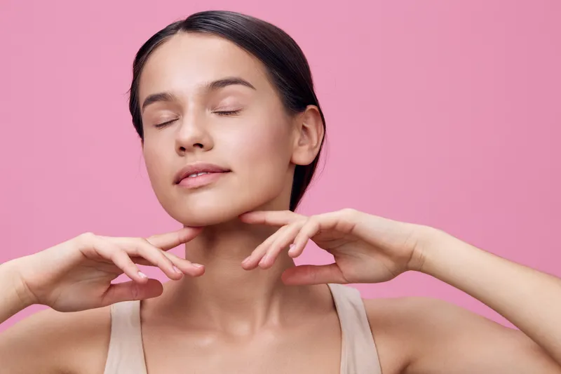 Young woman enjoying skincare routine with closed eyes, soft expression, neutral outfit, against a pink background, promoting self care and wellness