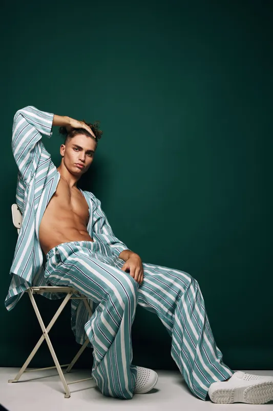 Young man in striped pajamas sitting on chair with hand on head against dark green background, casual pose and relaxed mood in studio setting.