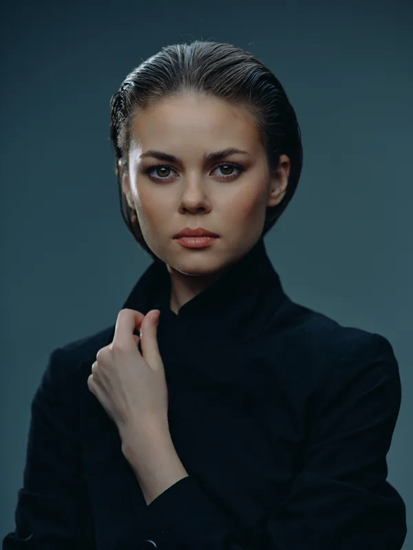 Serious woman with dark hair wearing black clothing against a dark background, showing confidence and elegance.