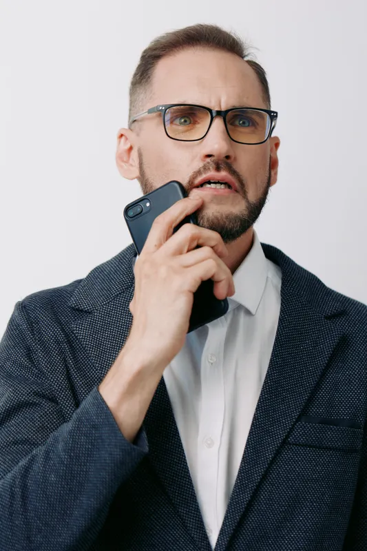 Professional male businessman on an isolated colored background, wearing glasses and a blazer, engaged in a phone call with a focused and anxious expression.