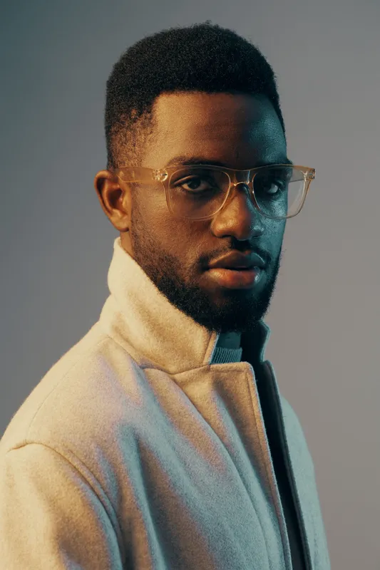 Stylish young Black man with glasses in a soft pastel background, showcasing modern fashion and confidence His unique hairstyle and expression reflect urban aesthetics