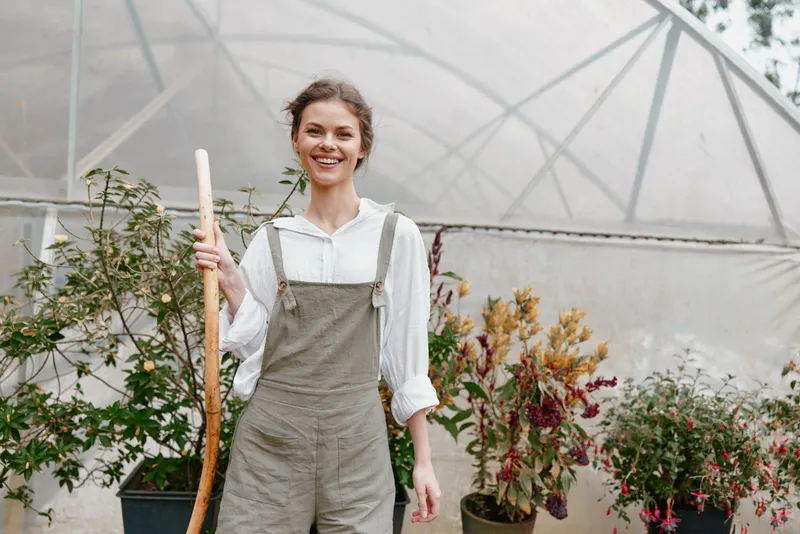 Young woman gardening in a greenhouse, smiling cheerfully while holding a gardening tool, surrounded by various plants and flowers, showcasing a joyful and sustainable lifestyle